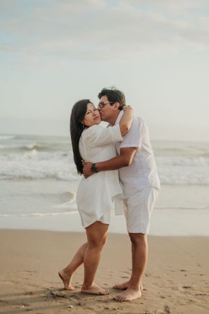 Couple in white outfits sharing a tender moment on a sandy beach.