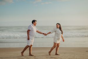 Couple in matching white outfits walking hand in hand along the shoreline.