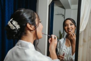 Reflection of someone applying makeup in a mirror while wearing a white top and hair accessory before a wedding.