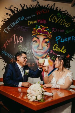 Wedding couple shares drinks at a table with colorful graffiti wall backdrop.