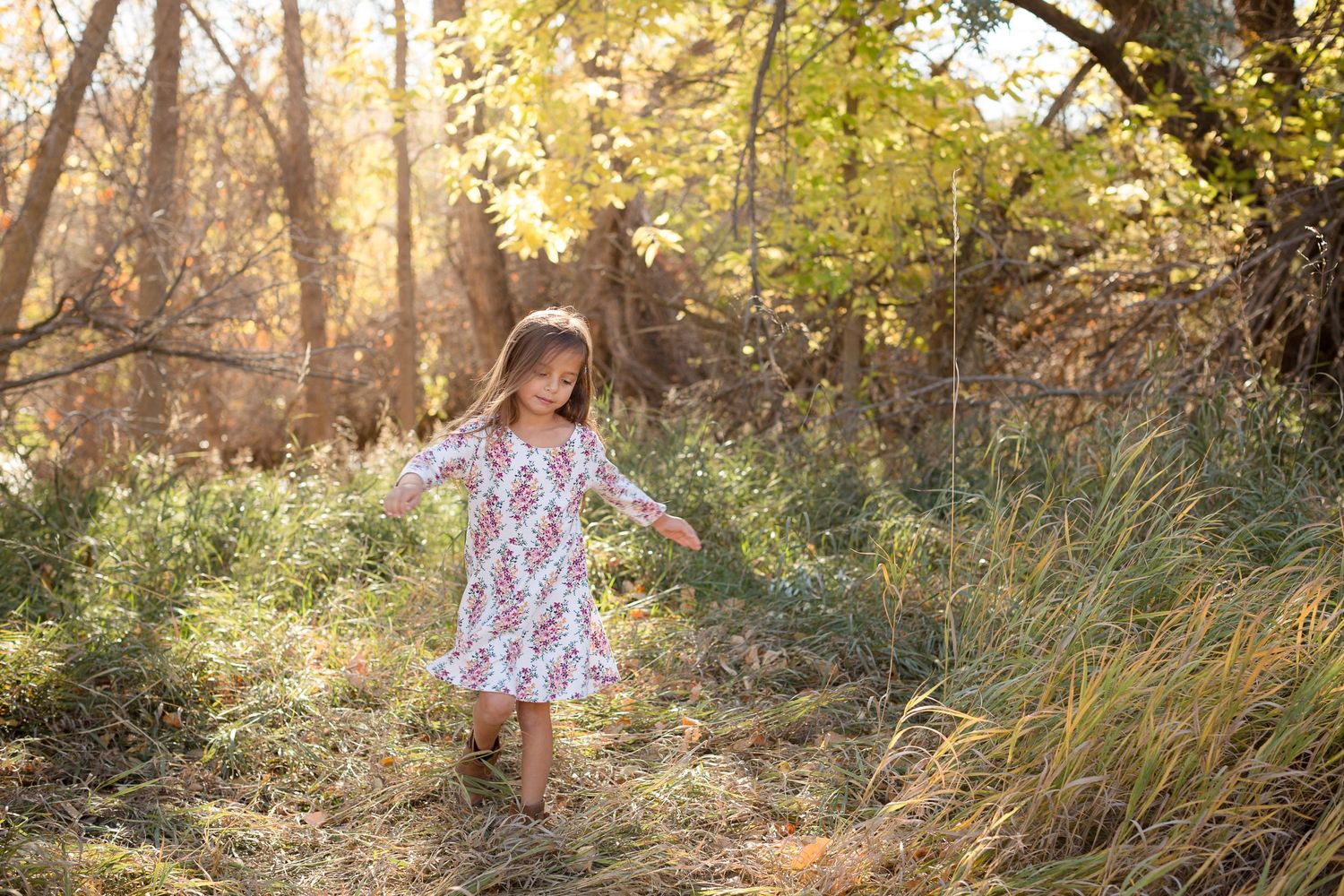A young child in a floral dress twirling through tall grass during golden hour.