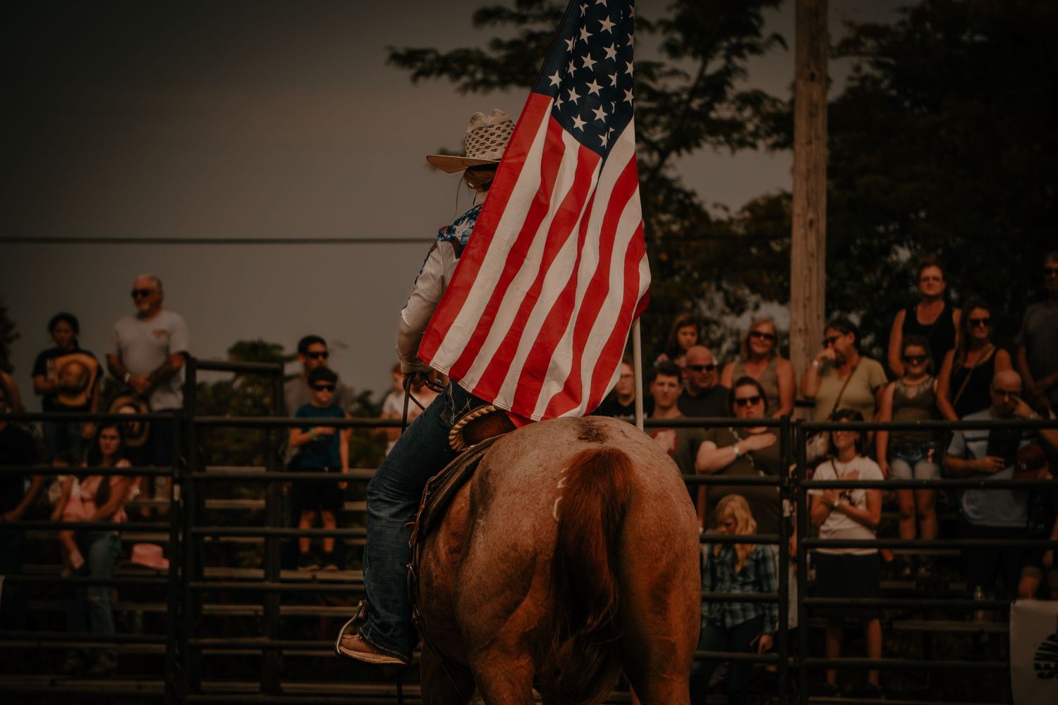 Horseback rider holding American flag with spectators in bleachers at dusk.