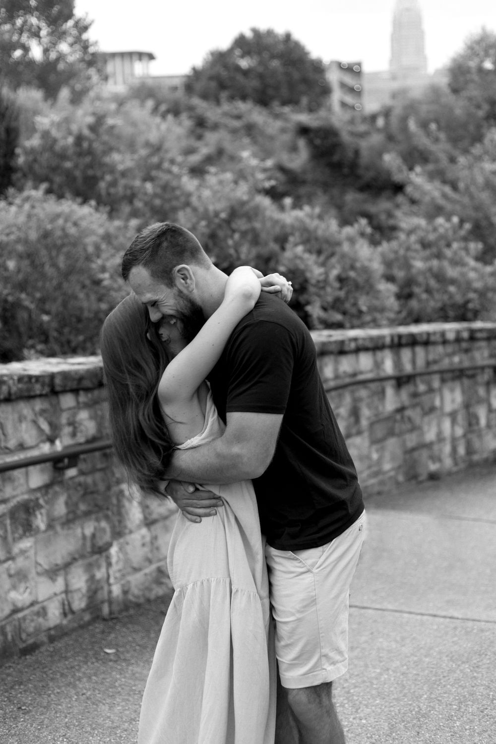 A woman throws her arms around her fiancé's neck in front of the Charlotte skyline. Black and white.