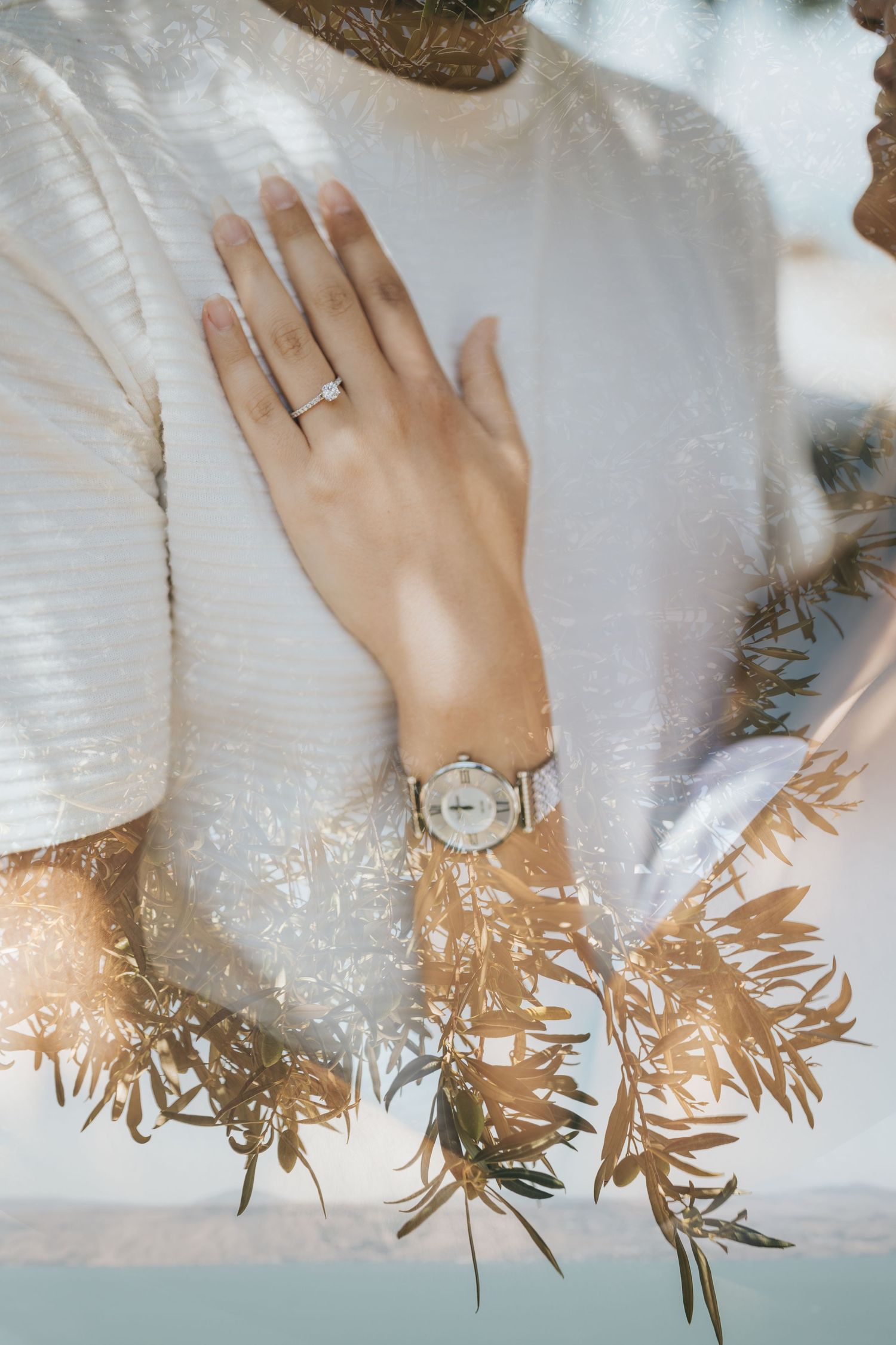 Close-up of engagement ring on hand with watch against white fabric and leafy shadows.
