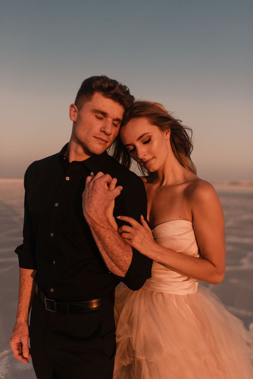 A romantic couple poses in formal attire on a salt flat at dusk.