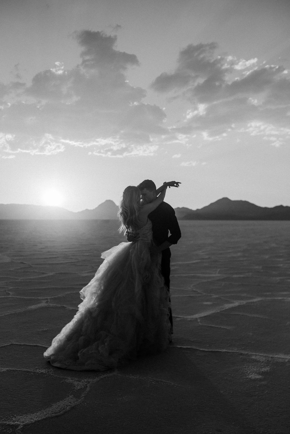 Silhouetted couple embracing at sunset on salt flats with mountains in background.