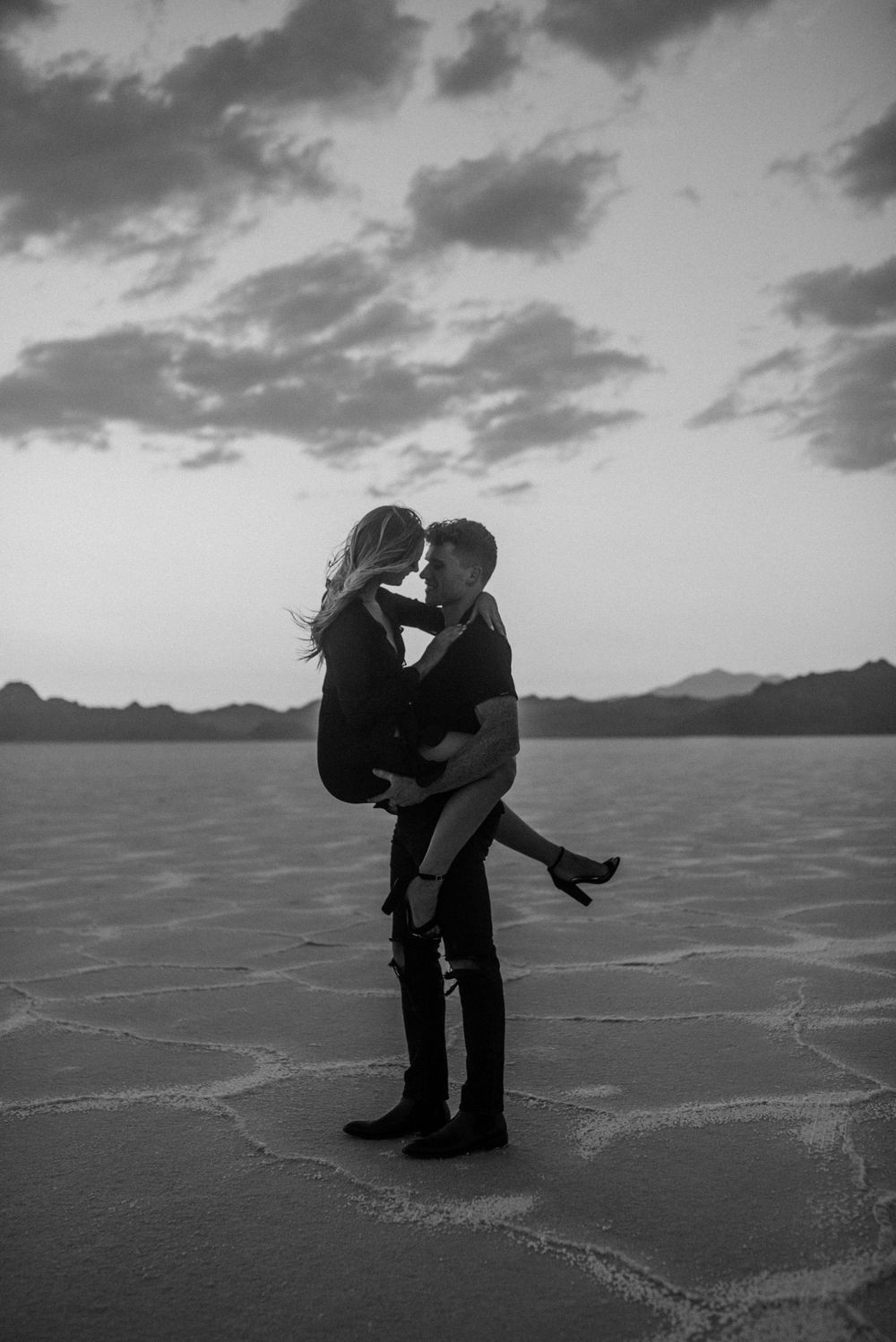 A black and white photo of a couple embracing on a salt flat with cloudy sky.