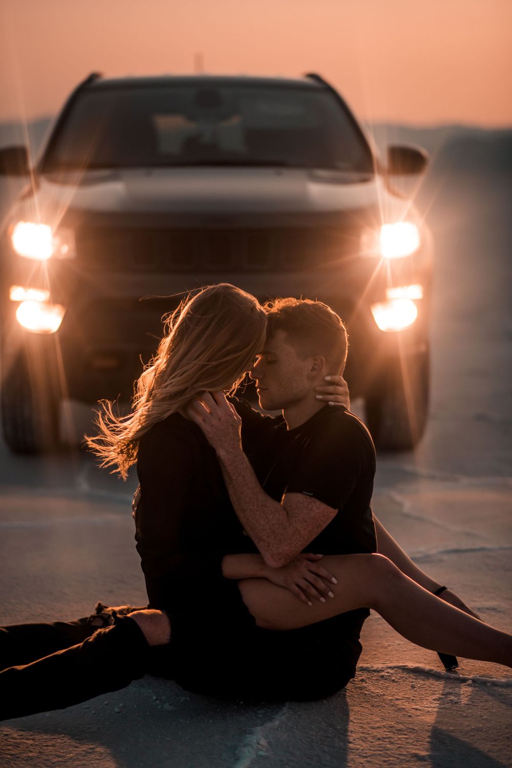 A couple shares an intimate moment in front of car headlights at sunset.