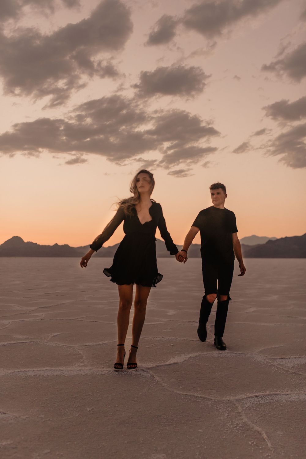 A couple walks hand in hand across a salt flat at sunset with dramatic clouds overhead.