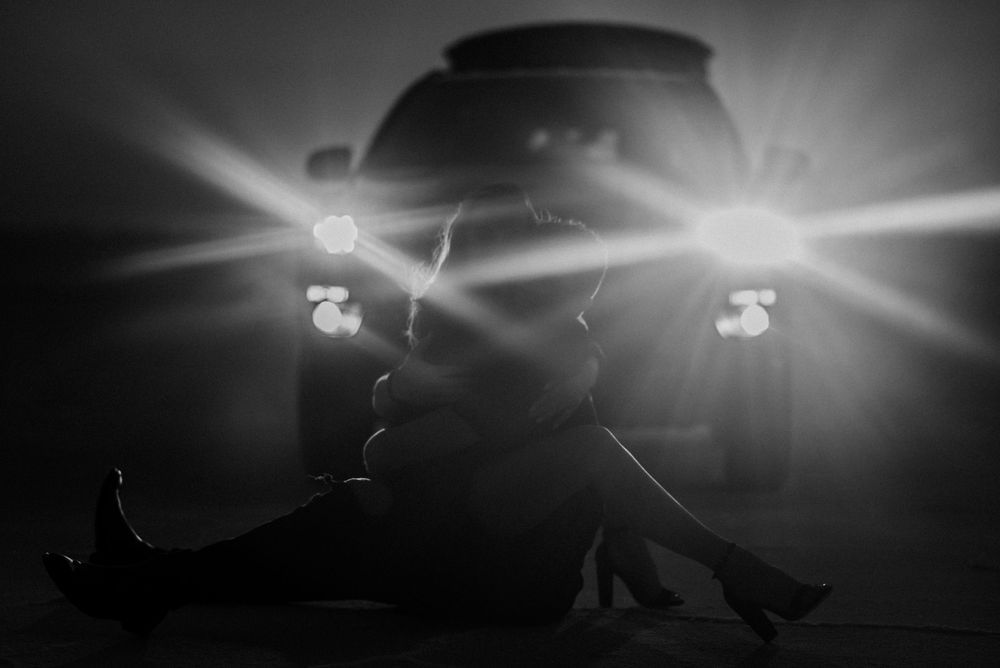 Silhouetted couple embracing in front of car headlights creating dramatic lighting effect.