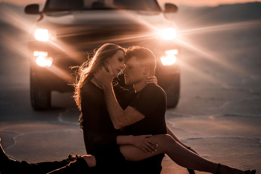 A romantic couple embraces in front of car headlights at dusk on a salt flat.