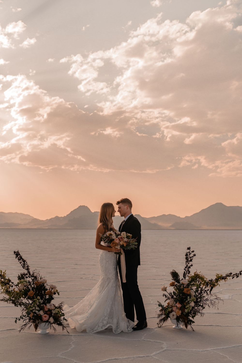 Wedding couple standing between floral arrangements on salt flats at sunset.