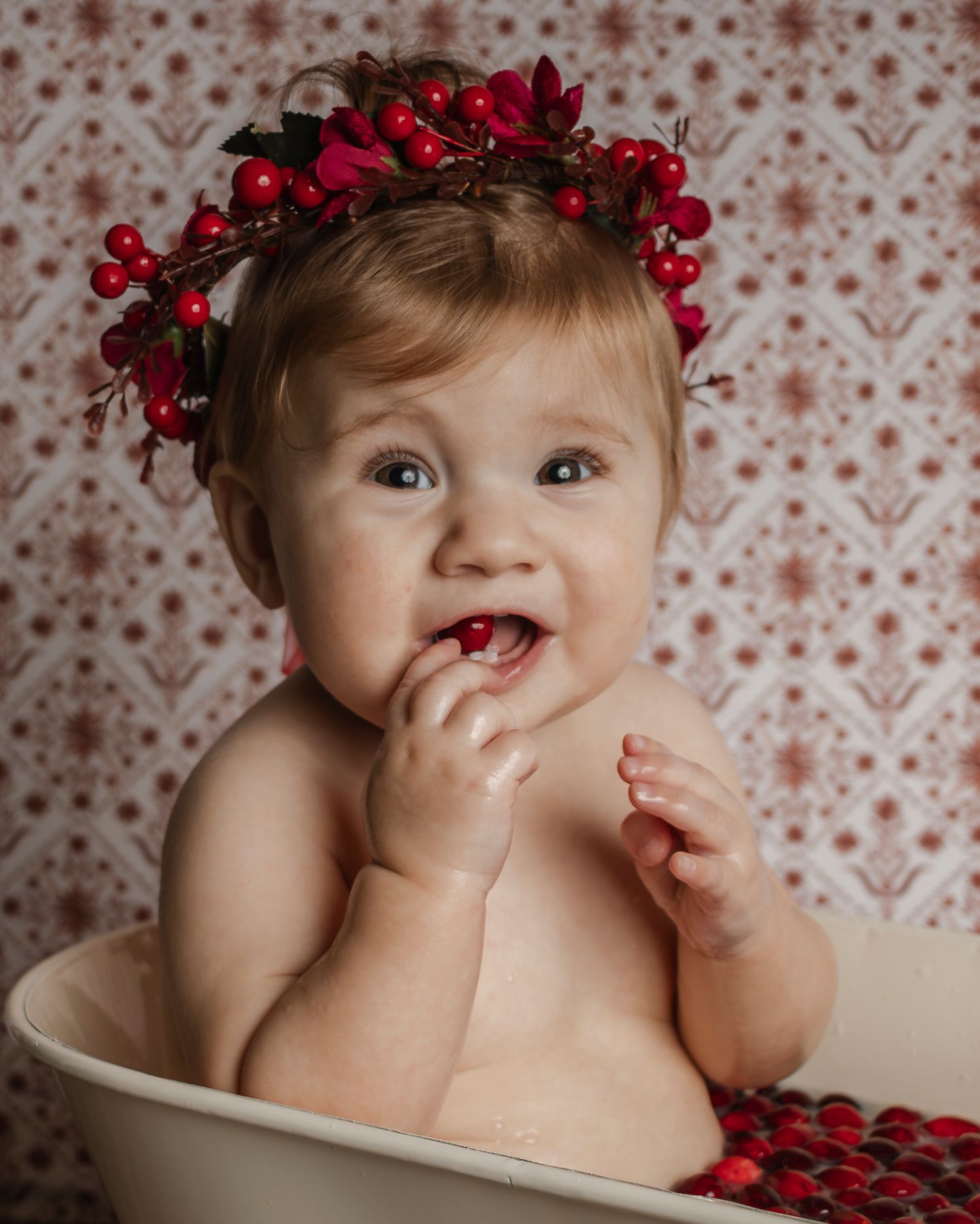 A baby girl in a miniature bathtub full of milk and cranberries puts a berry in her mouth.