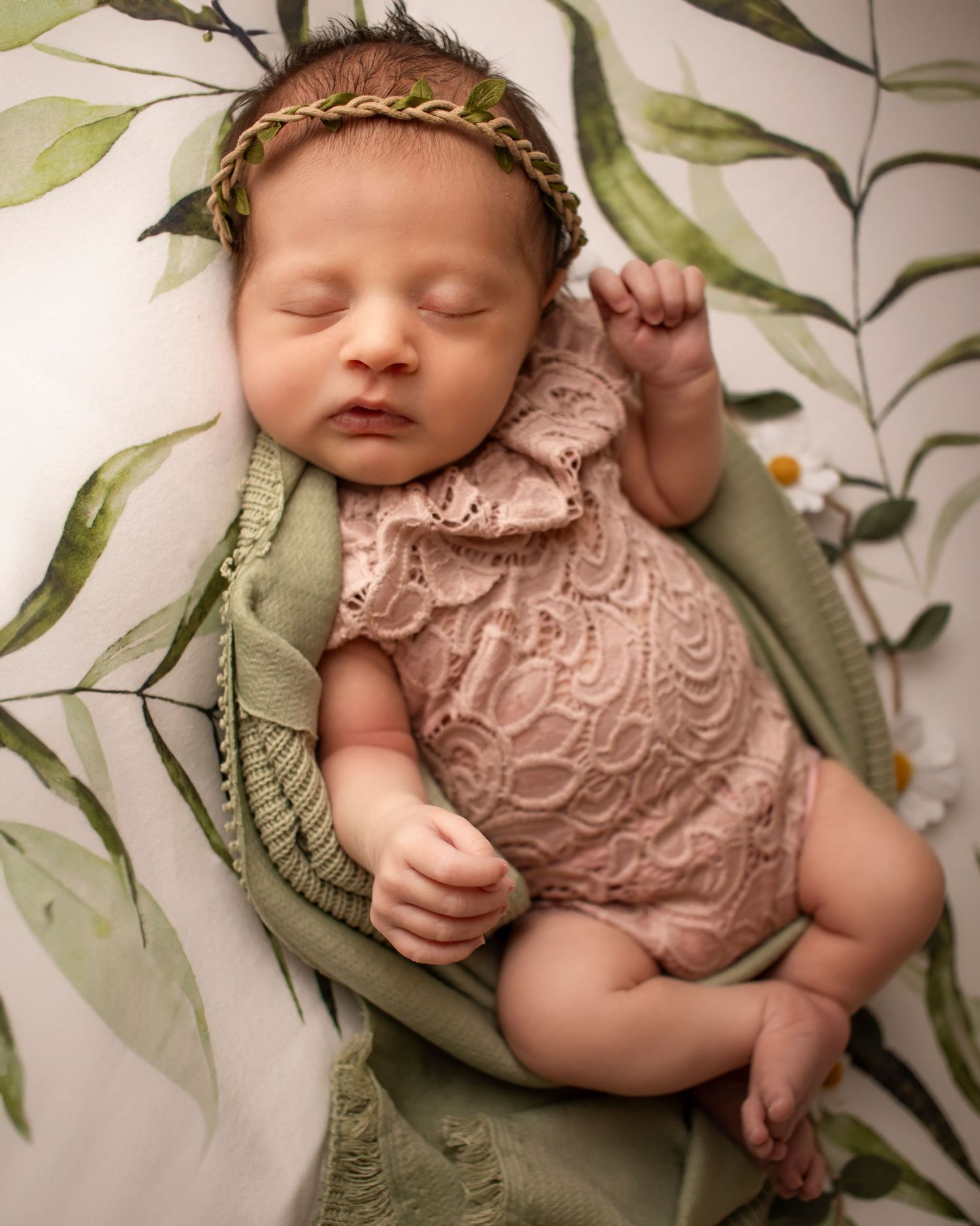 A newborn girl is curled across a botanical backdrop. She is sleeping peacefully and wearing a blush lace romper.