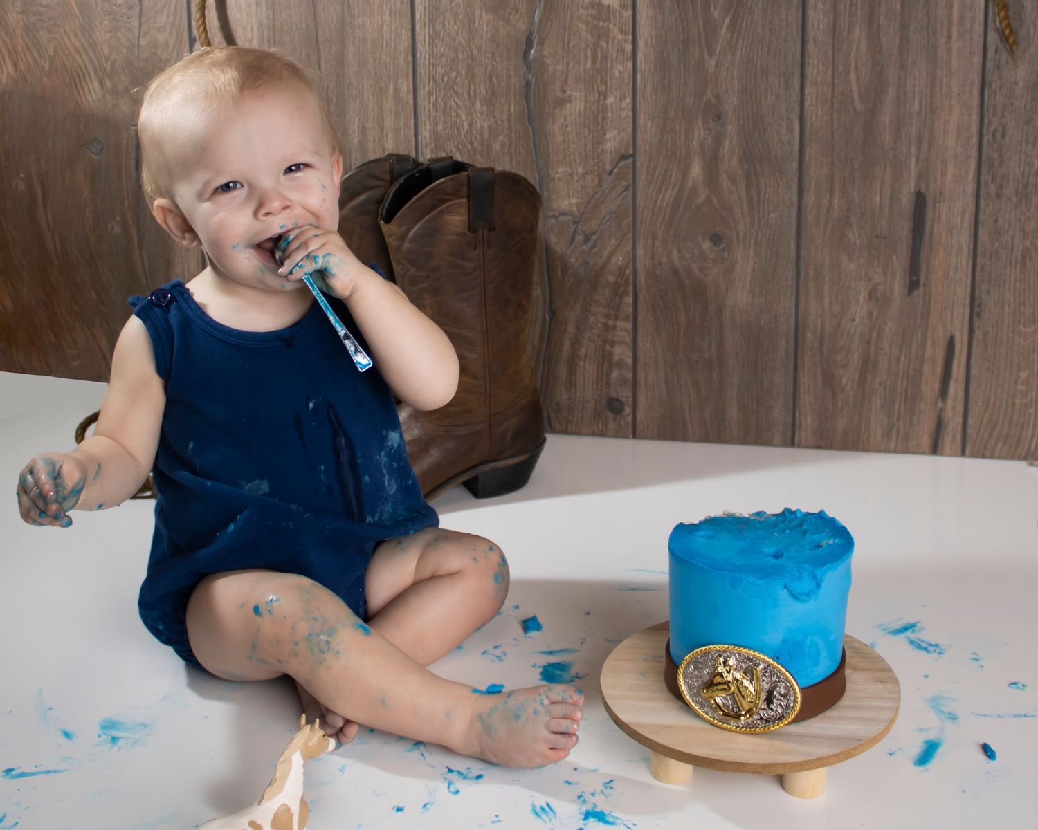 A baby boy uses a silver spoon to eat bright blue frosting off a western style smash cake. Frosting is all over the place