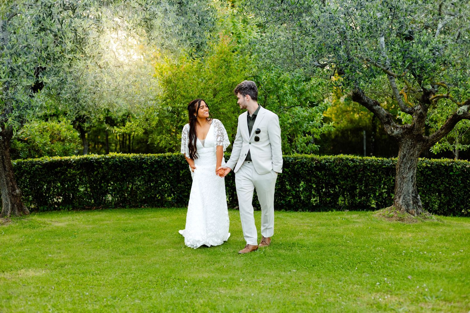 A couple in white attire shares a romantic moment in a sunlit garden setting.