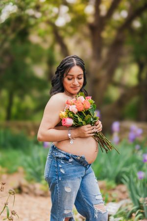 A pregnant woman in distressed jeans holds a bouquet of peach and coral roses in a garden setting.