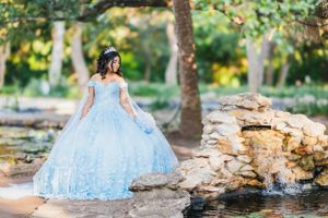 A light blue quinceañera ball gown with lace details sparkles near a stone waterfall in a sunlit garden setting.