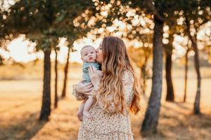 A parent holds a baby in a sun-drenched forest setting during golden hour with trees silhouetted in the background.