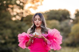Someone in a vibrant pink dress with dramatic ruffled sleeves blows out birthday candles at sunset in an outdoor setting.