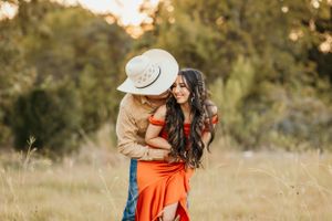 A couple embraces in a golden field at sunset with one wearing an orange dress and the other in a cowboy hat.