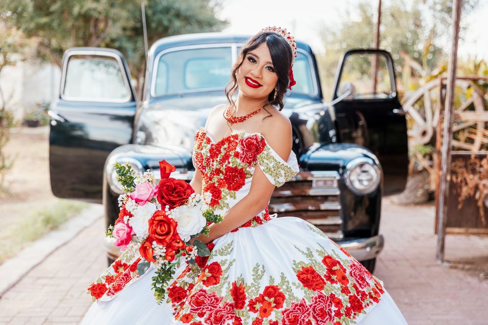 A person in a white and red floral quinceañera dress poses with a bouquet in front of a vintage black truck.