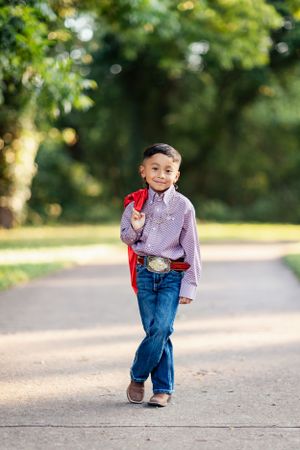 Young child in purple shirt and jeans walks confidently on path with red gown against blurred green background.