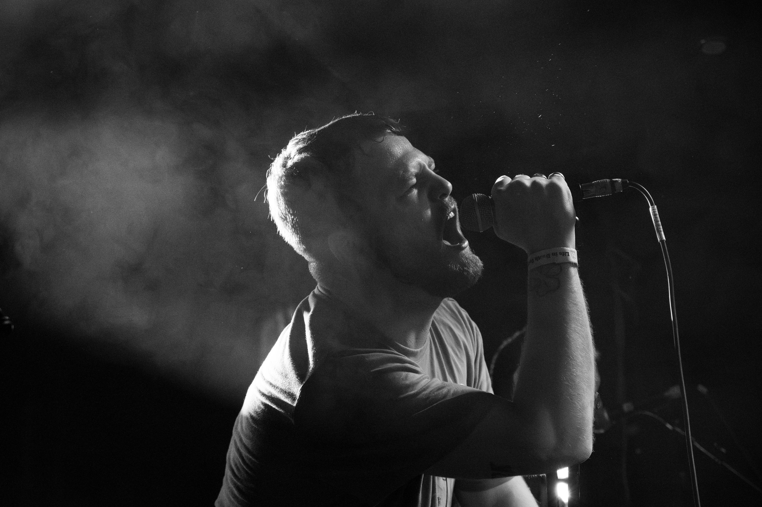 Dramatic black and white concert photo of performer singing into microphone with stage lighting.