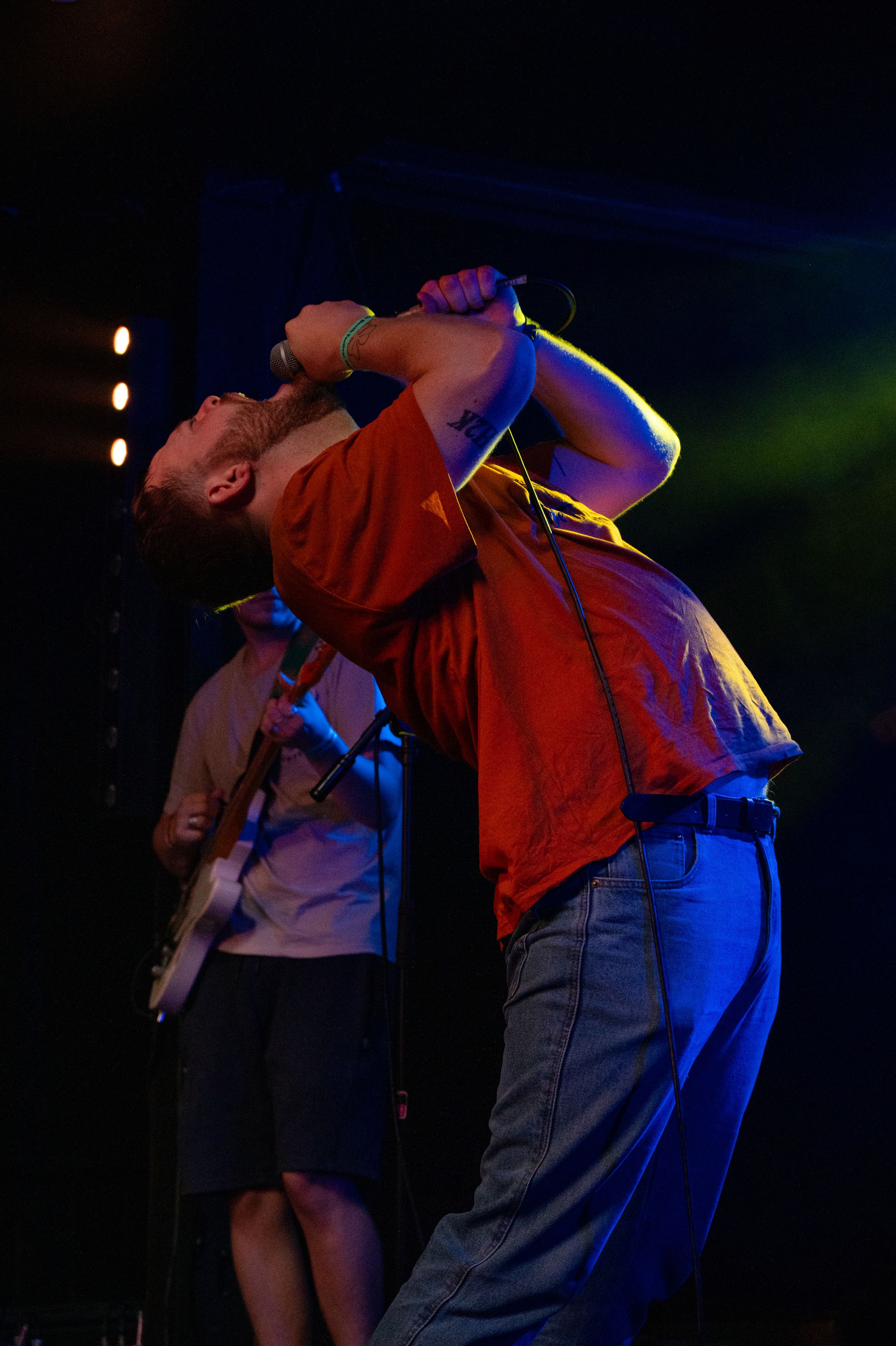 Performer in red shirt leans back dramatically while singing on stage under colorful spotlights.