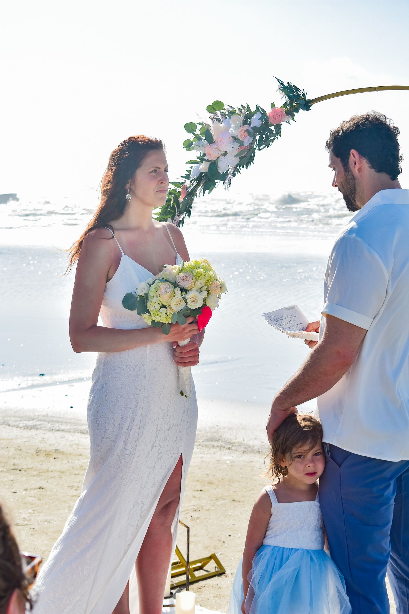 A beach wedding ceremony with a floral arch, white dress, and yellow bouquet on a sunny oceanfront.