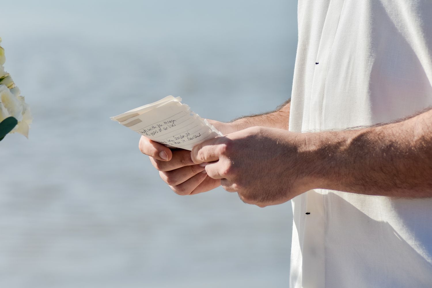 Hands holding wedding ceremony papers against blurred ocean background.