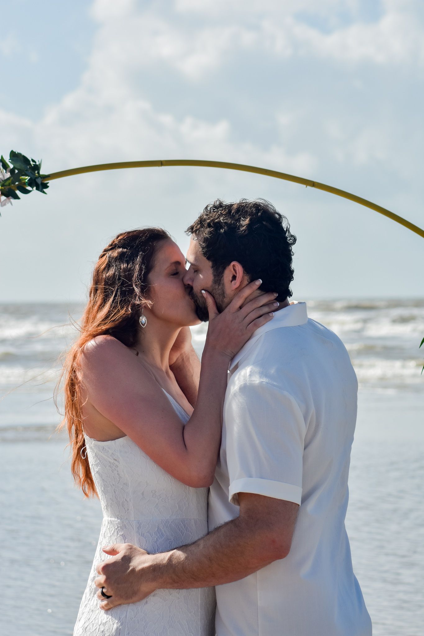 A couple shares a romantic kiss on a sunny beach with the ocean waves in the background.