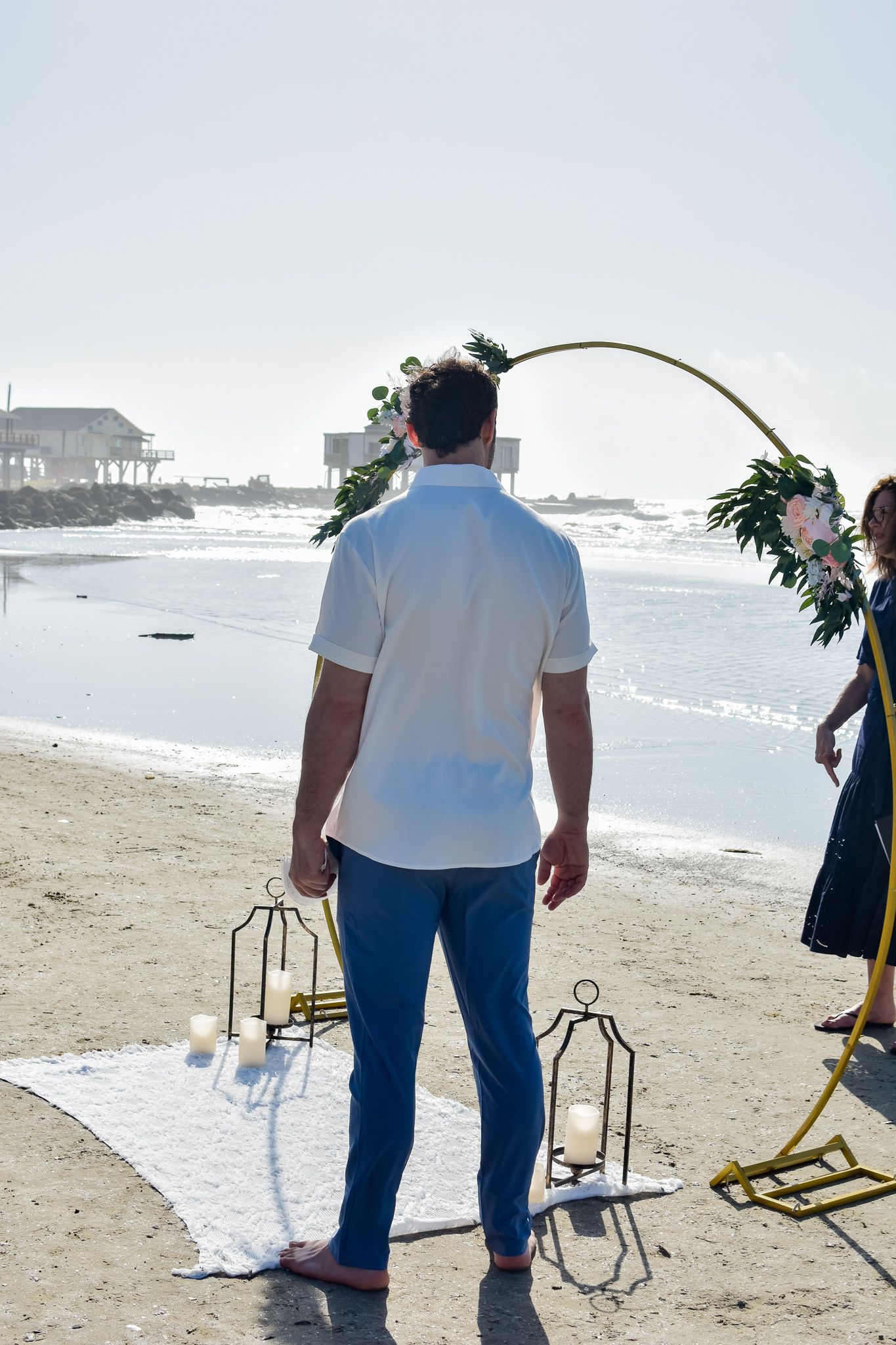 Gold wedding arch with floral decorations and lanterns set up on beach with ocean view.