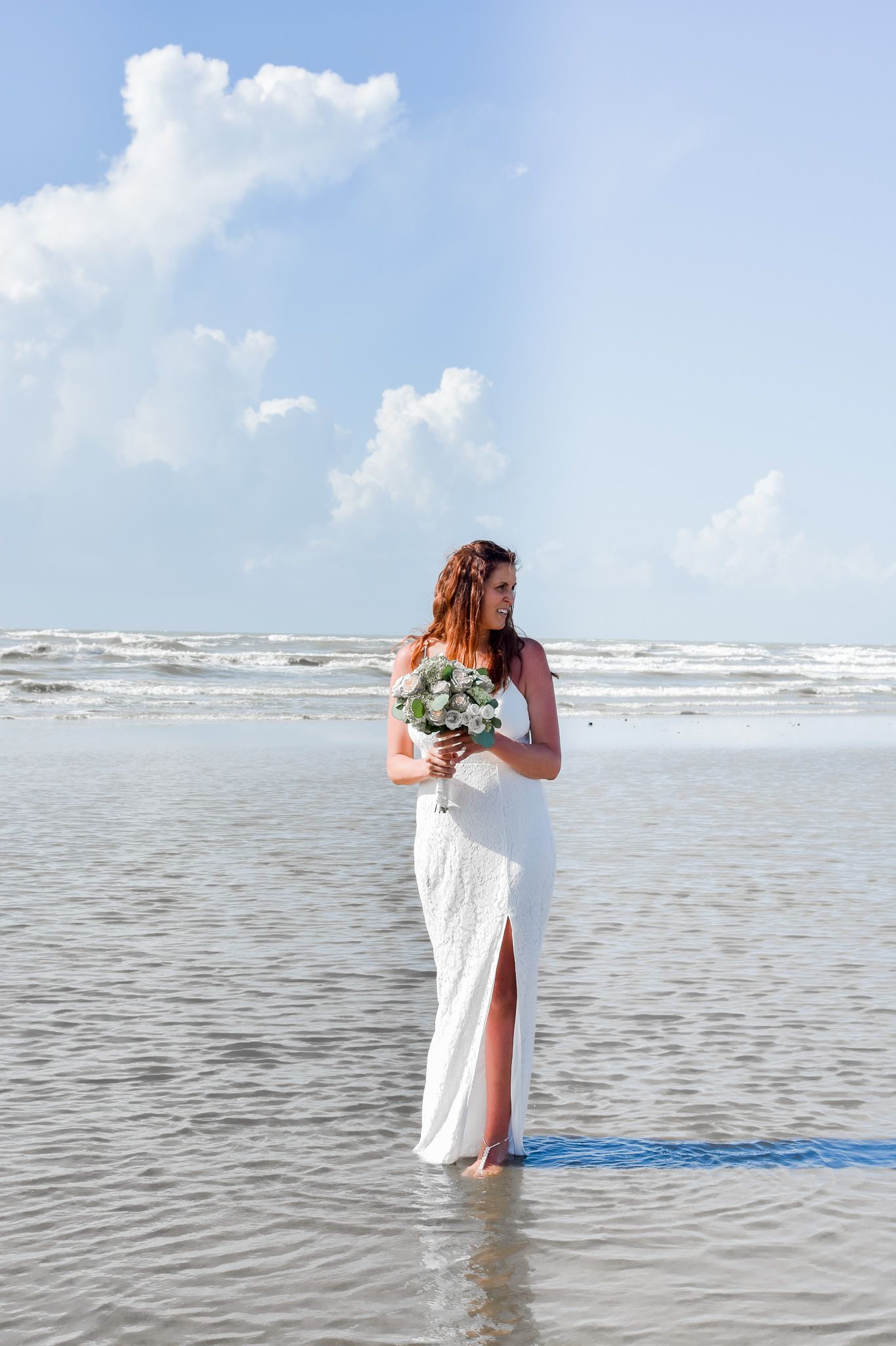 Bride in flowing white dress stands alone on beach holding bouquet.