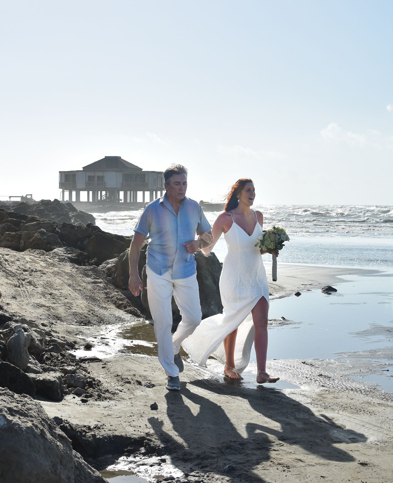 Couple walking along rocky beach shoreline with stilt house visible in background.