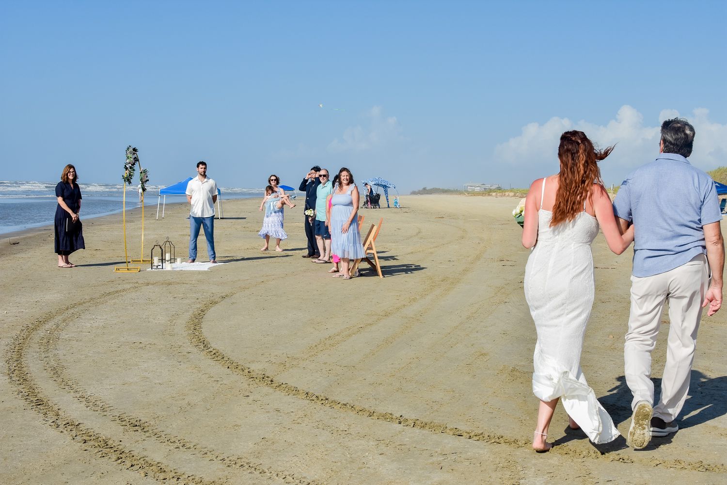 Wedding party walking along beach with tire tracks in sand and stilt houses in background.