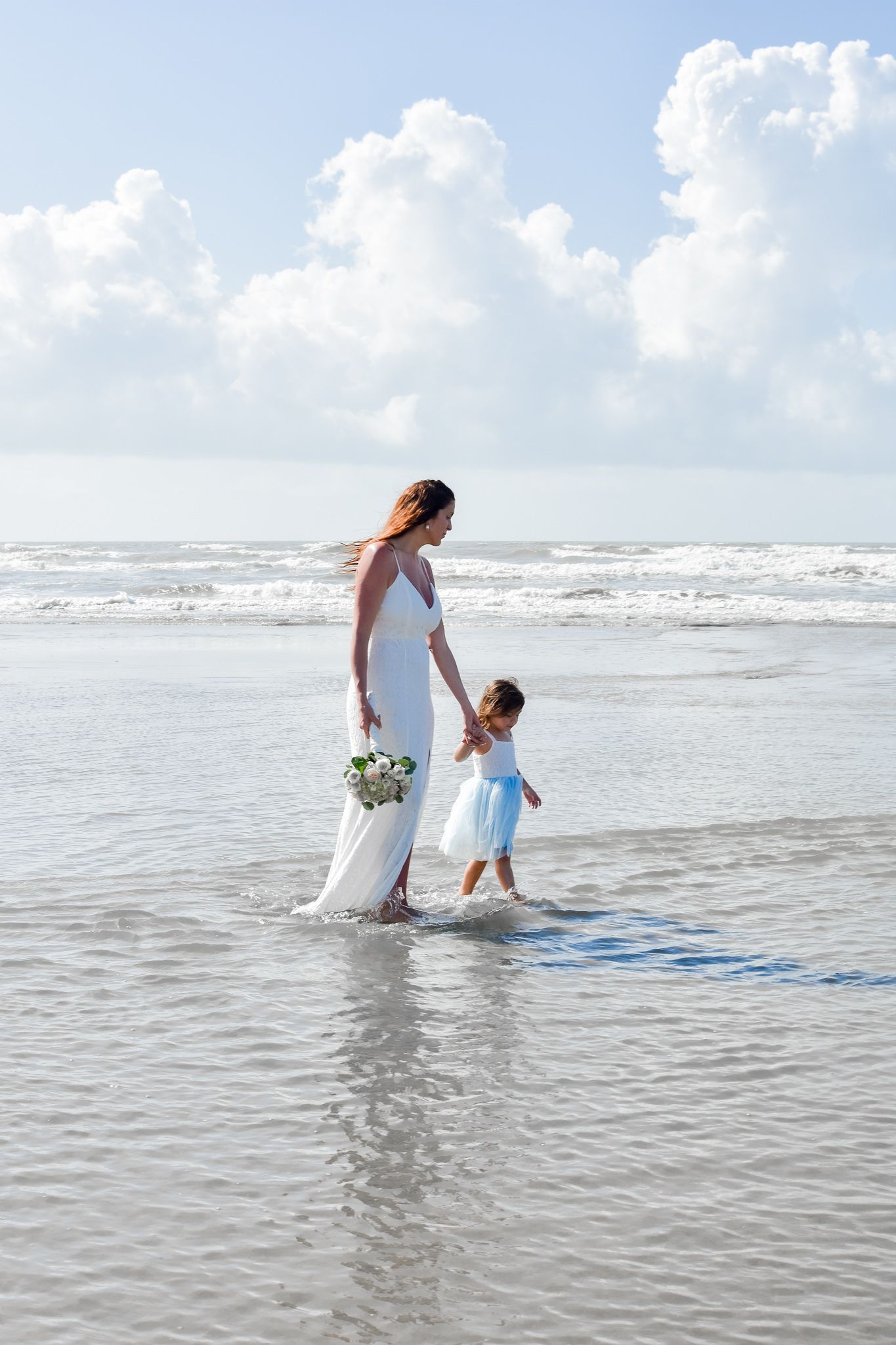 Woman in white dress walking with small child along ocean shoreline.