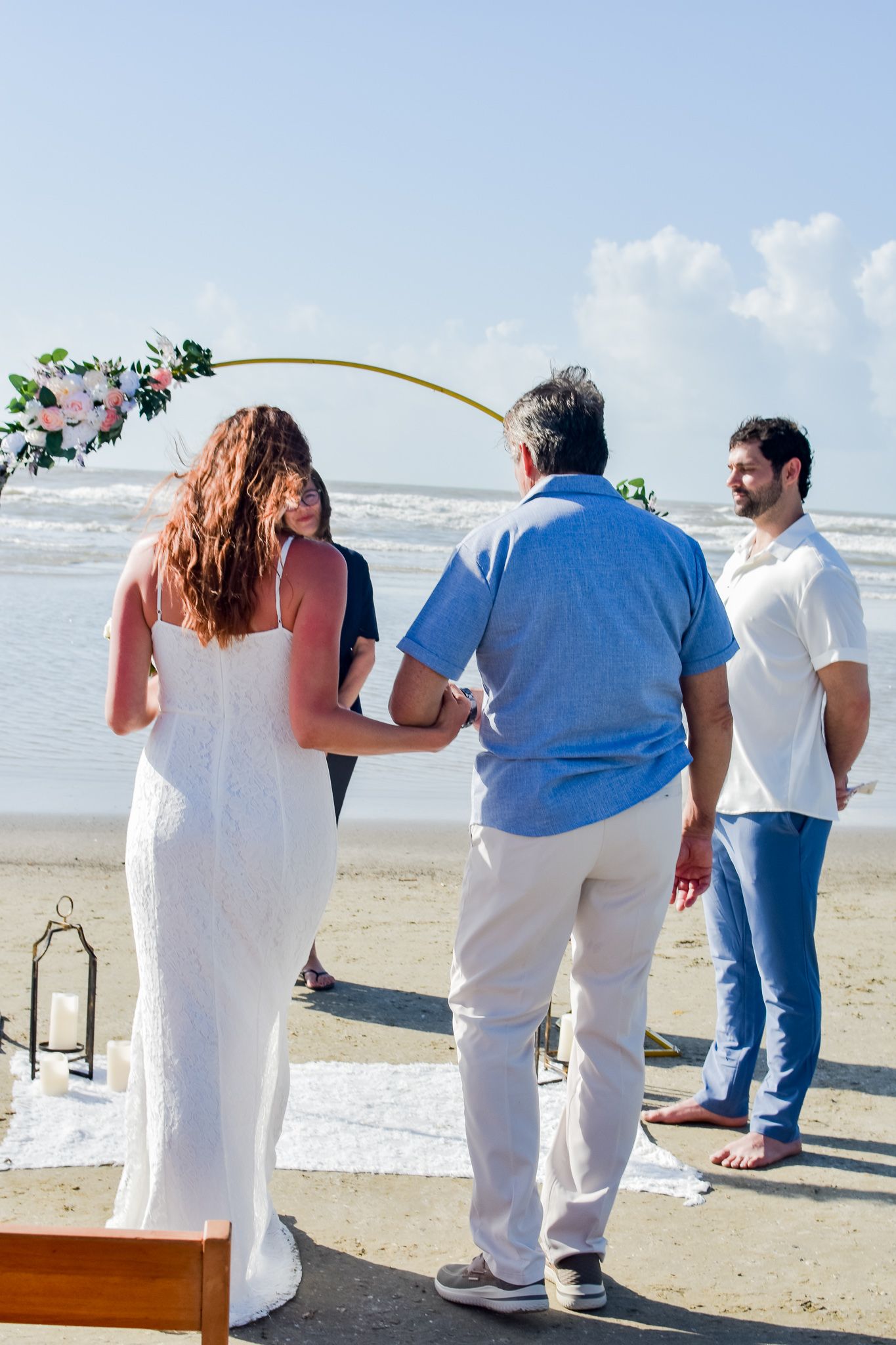 Beach wedding ceremony with decorative gold arch and small gathering against ocean backdrop.