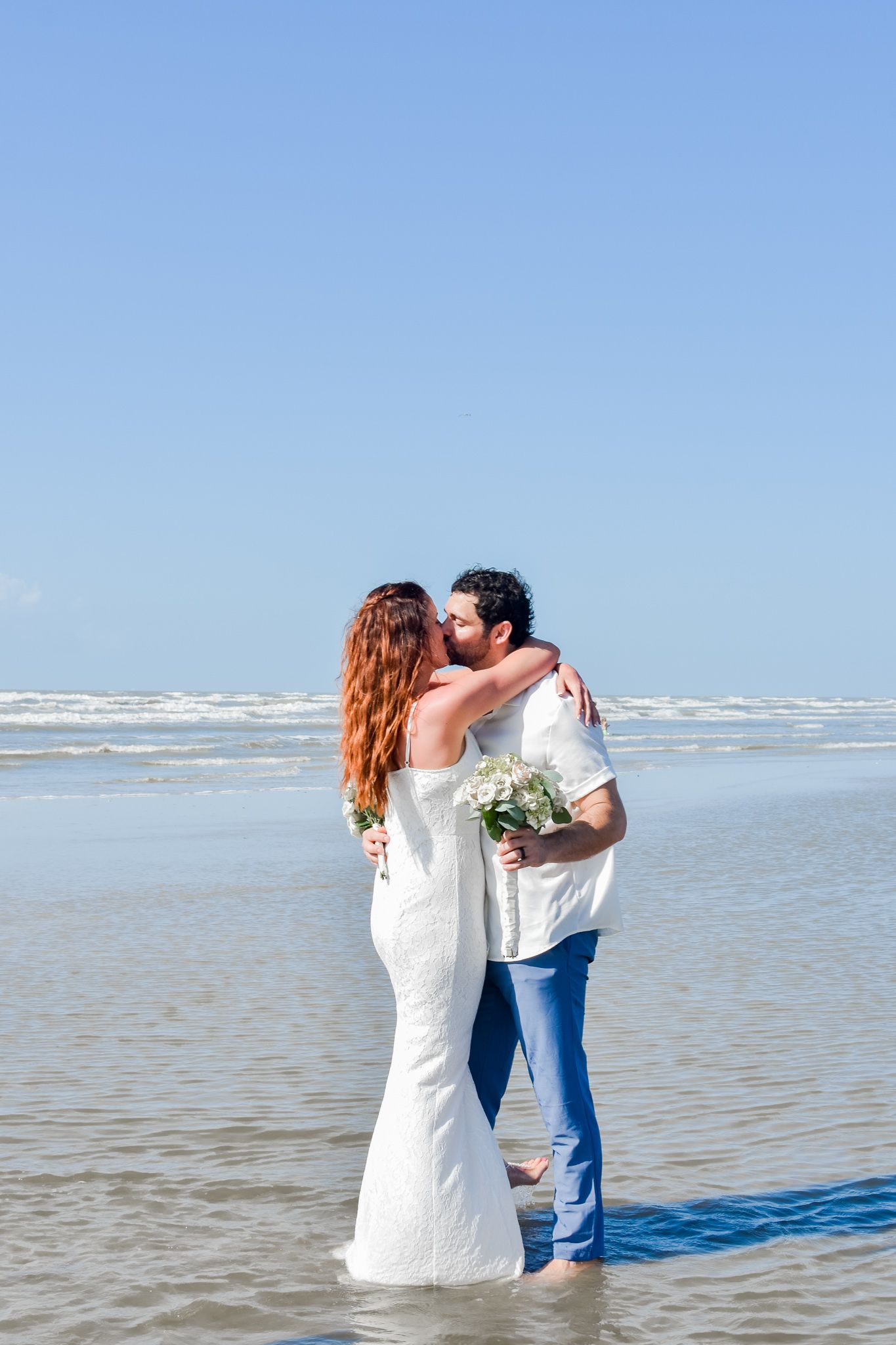 Romantic couple in wedding attire embracing in shallow ocean water.