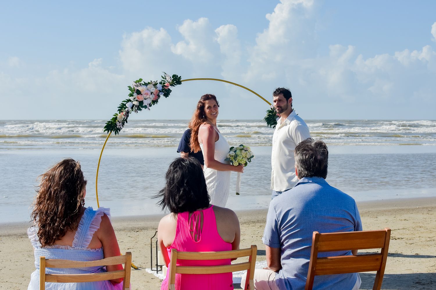 Intimate beach wedding ceremony under a floral arch with guests seated on wooden chairs by the ocean.