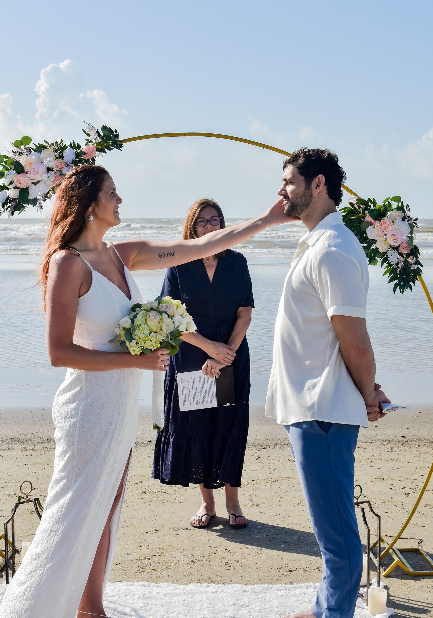 Intimate beach wedding ceremony with officiant under curved floral arch by the ocean.
