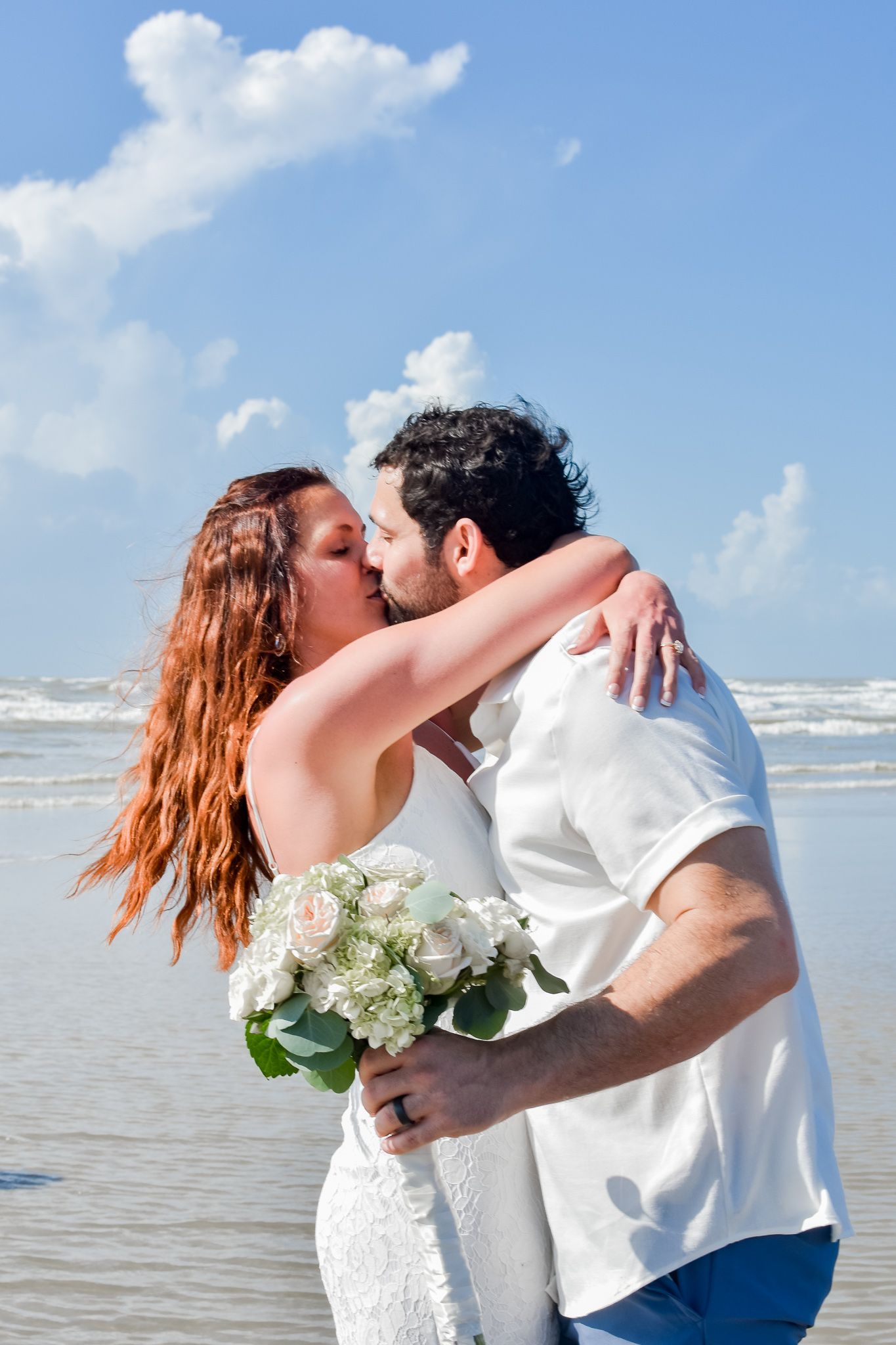 Newlyweds embrace and kiss on beach with white flower bouquet.
