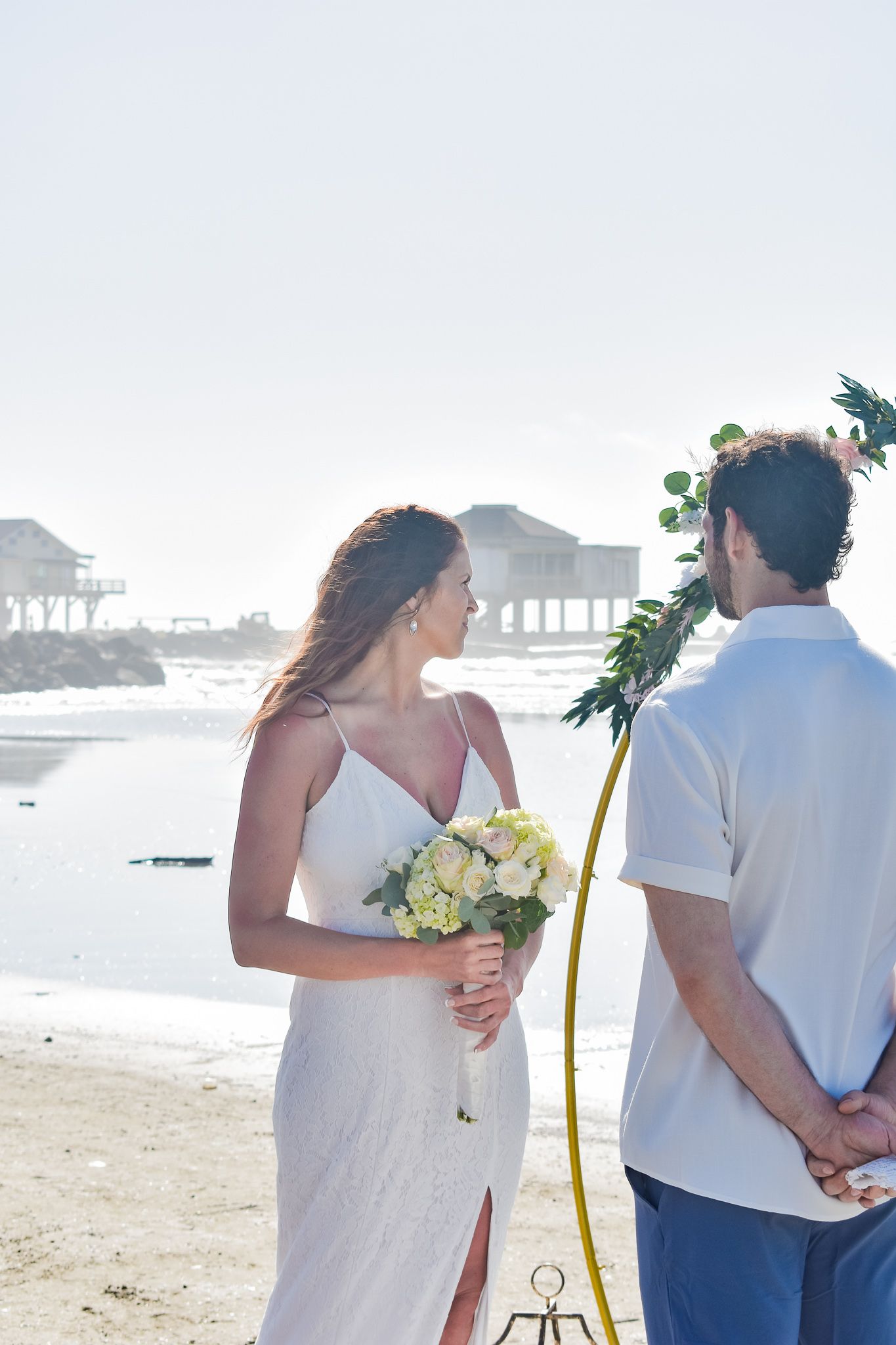 Beachside wedding with pier houses visible in misty background and floral arch decoration.