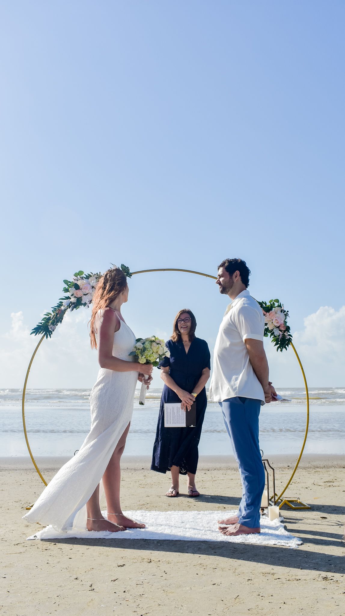Minimalist beach wedding ceremony under round golden arch with officiant and white carpet.