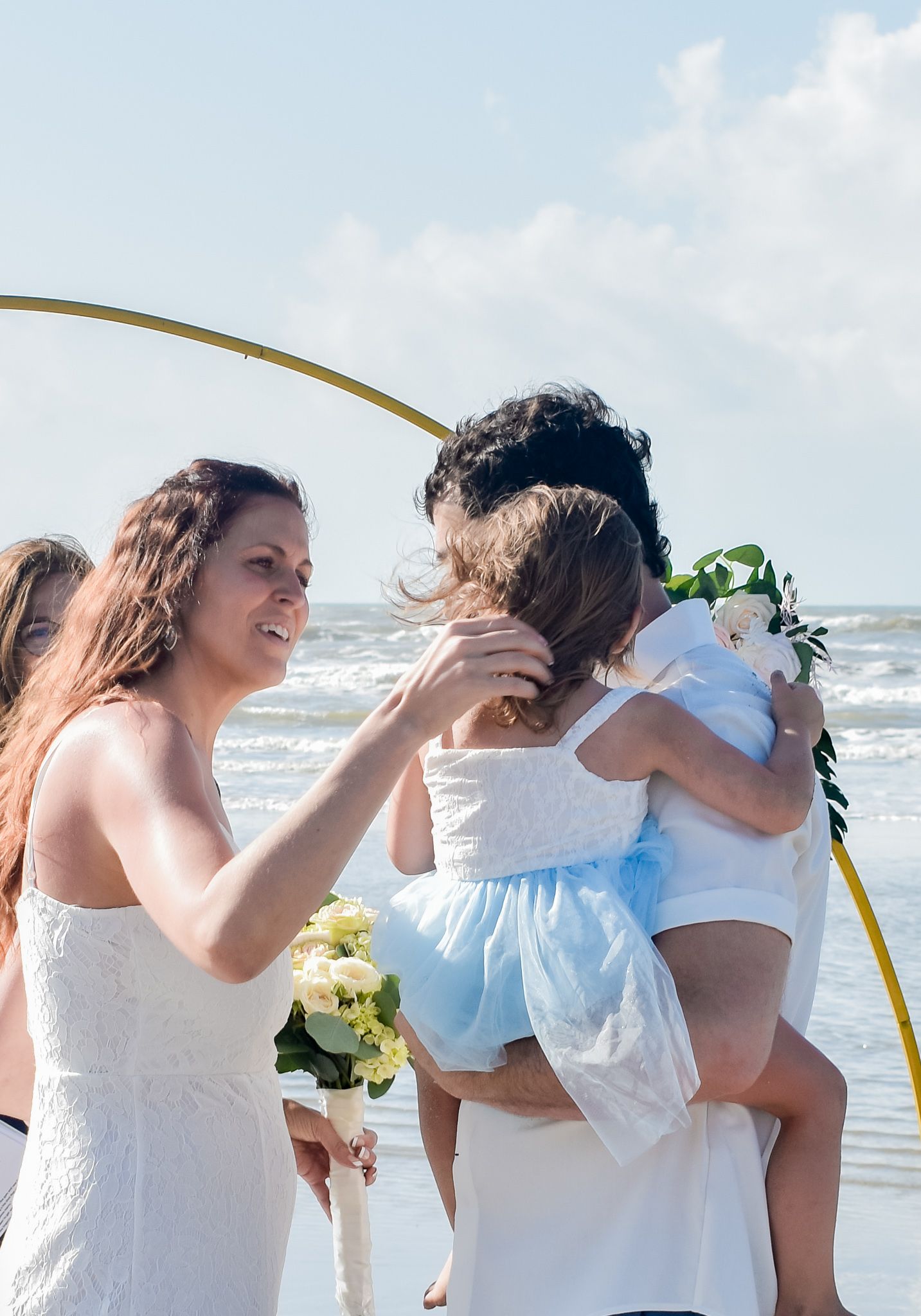 Emotional beach wedding moment under golden arch with ocean waves in background.