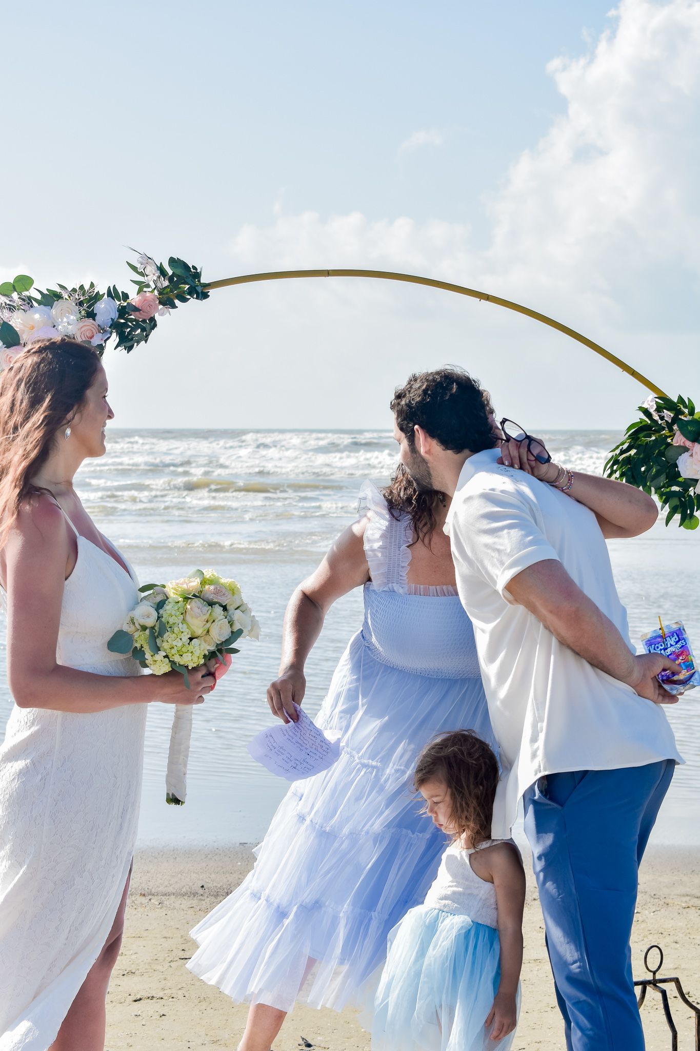 Casual beach wedding ceremony with bride holding yellow bouquet under curved floral arch.