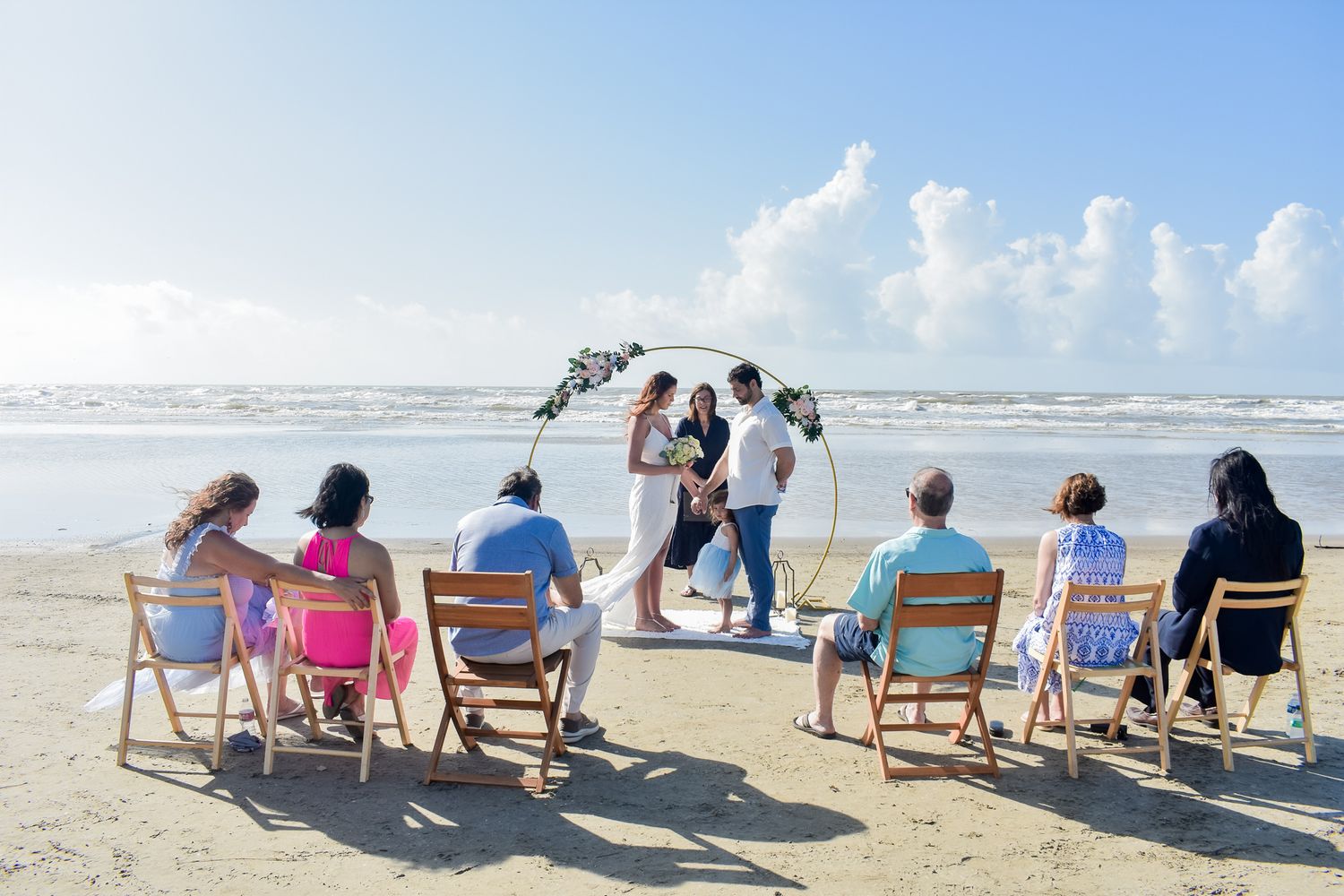 Beach wedding ceremony with guests seated in chairs watching couple under floral arch by the ocean.