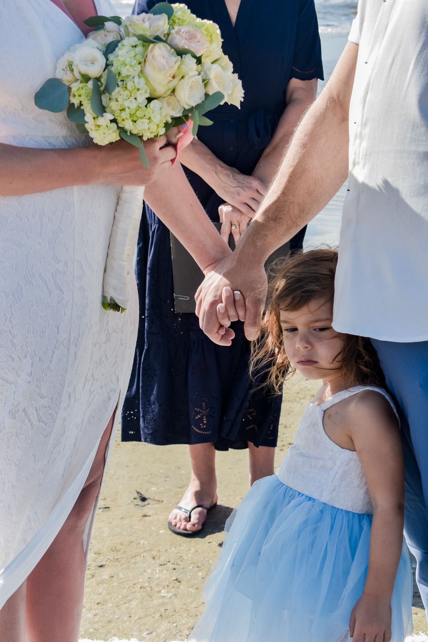An intimate beach wedding moment showing joined hands and a bouquet of white and yellow flowers.