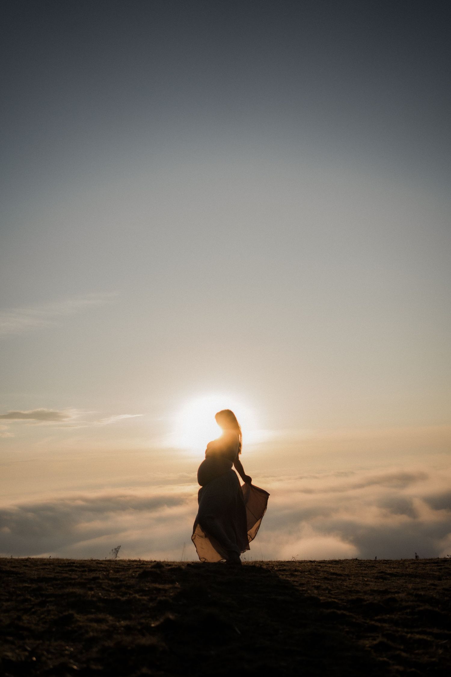 Silhouette of a figure in a flowing dress against a dramatic sunset sky above clouds.