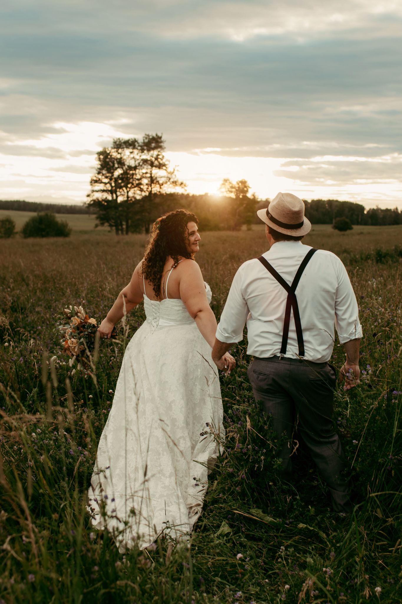 A couple walks through a golden field at sunset with suspenders and a flowing white dress.